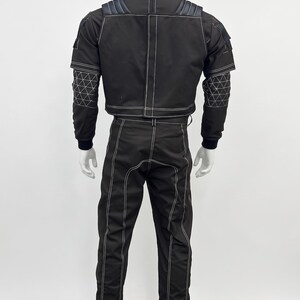 May include: A black two-piece outfit consisting of a high-neck jacket and matching trousers. The jacket features shoulder padding, long sleeves with a white geometric grid pattern on the forearms, and white contrast stitching throughout.