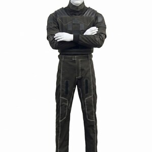 May include: A dark olive green tactical-style outfit consisting of a long-sleeved top with a high collar, shoulder padding, and matching trousers. The garment features prominent white contrast stitching, rectangular black patches, and a belt.