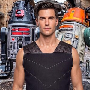 May include: A sleeveless black vest featuring a textured dark brown shoulder panel and white stitched chevron detailing on the front. The garment is worn in front of two robotic figures, one black and red and the other orange and white.