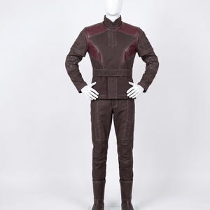 May include: A dark brown long-sleeved jacket and matching trousers set on a mannequin. The jacket features a high collar, a wide waist belt, and dark red shoulder panels. The sleeves and pants have white topstitching and geometric patterns.