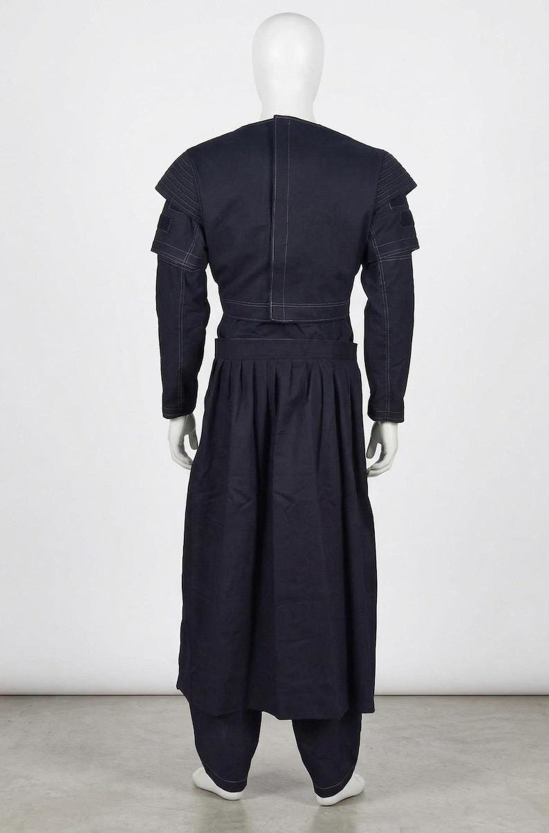 May include: A dark navy blue ensemble consisting of a cropped jacket with structured shoulders and long sleeves, paired with a high-waisted, floor-length pleated skirt. The garment features visible white contrast stitching throughout the design.