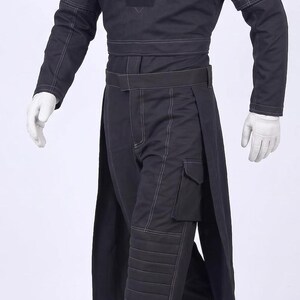 May include: A black tactical-style outfit consisting of a long-sleeved top with white contrast stitching and a central patch, paired with matching trousers featuring a side cargo pocket, quilted knee panels, and an integrated skirt-like overlay.