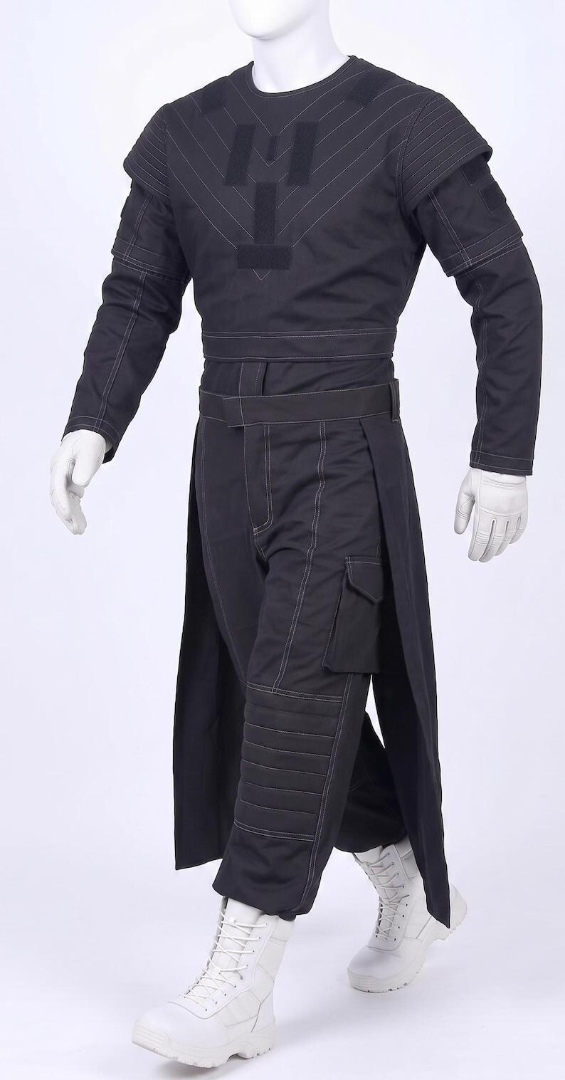 May include: A black tactical-style outfit consisting of a long-sleeved top with white contrast stitching and a central patch, paired with matching trousers featuring a side cargo pocket, quilted knee panels, and an integrated skirt-like overlay.