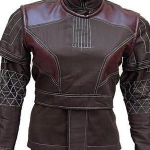 May include: A dark brown jacket featuring maroon shoulder panels, a high collar, and white topstitching. The sleeves display a geometric triangular pattern in white thread, while the bodice includes a horizontal waistband and vertical stitching.