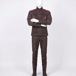 May include: A dark brown long-sleeved jacket and matching trousers set featuring a high collar, quilted sleeve detailing, and dark maroon shoulder panels. The outfit includes visible light-colored stitching throughout and dark brown boots.