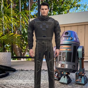 May include: A dark olive green tactical uniform featuring a high-neck top with structured shoulder panels and matching trousers with white contrast stitching, standing beside a metallic, iridescent blue and copper-toned cylindrical droid figure.