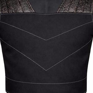 May include: A black vest featuring a square neckline and textured dark brown leather shoulder panels. The main body is made of a smooth black fabric with prominent light gray V-shaped stitching details across the chest and a horizontal seam at the waist.