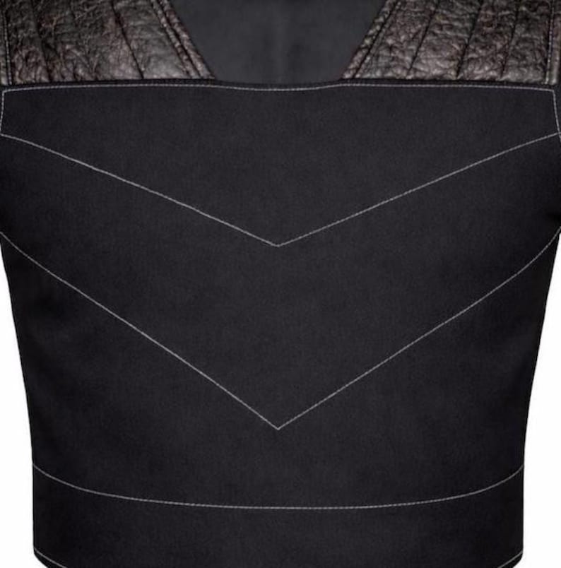 May include: A black vest featuring a square neckline and textured dark brown leather shoulder panels. The main body is made of a smooth black fabric with prominent light gray V-shaped stitching details across the chest and a horizontal seam at the waist.
