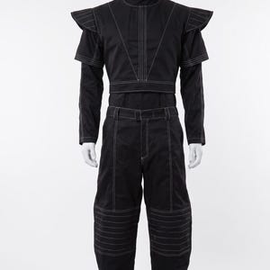 Pu&ograve; includere: Un completo nero composto da un top corto a maniche lunghe e pantaloni abbinati. Il top presenta un colletto alto, spalline strutturate e cuciture bianche a contrasto. I pantaloni includono passanti per cintura e cuciture orizzontali bianche.