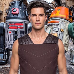 May include: A sleeveless brown tunic featuring a square neckline, textured shoulder panels, and subtle white chevron stitching across the chest. The garment is worn by a person standing between a black and silver droid and an orange and white droid.