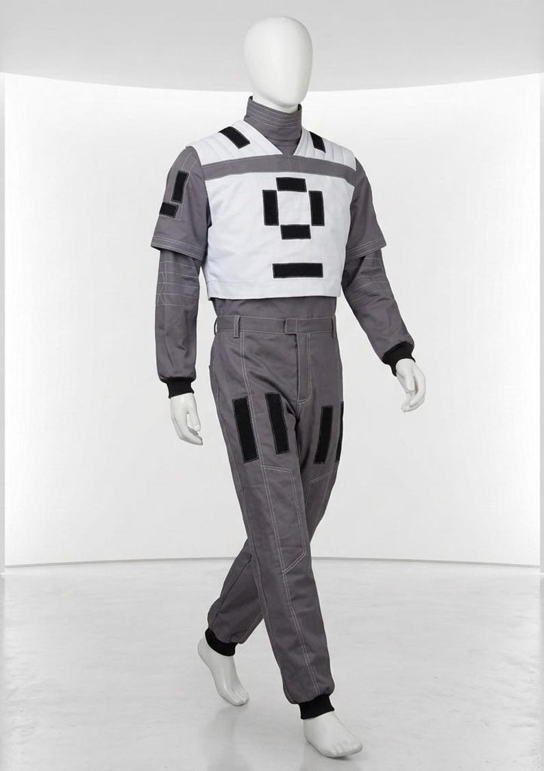 May include: A gray long-sleeved jumpsuit featuring a white chest overlay with black rectangular accents. The garment includes a high collar, black ribbed cuffs at the wrists and ankles, and additional black rectangular patches on the pants and sleeve.