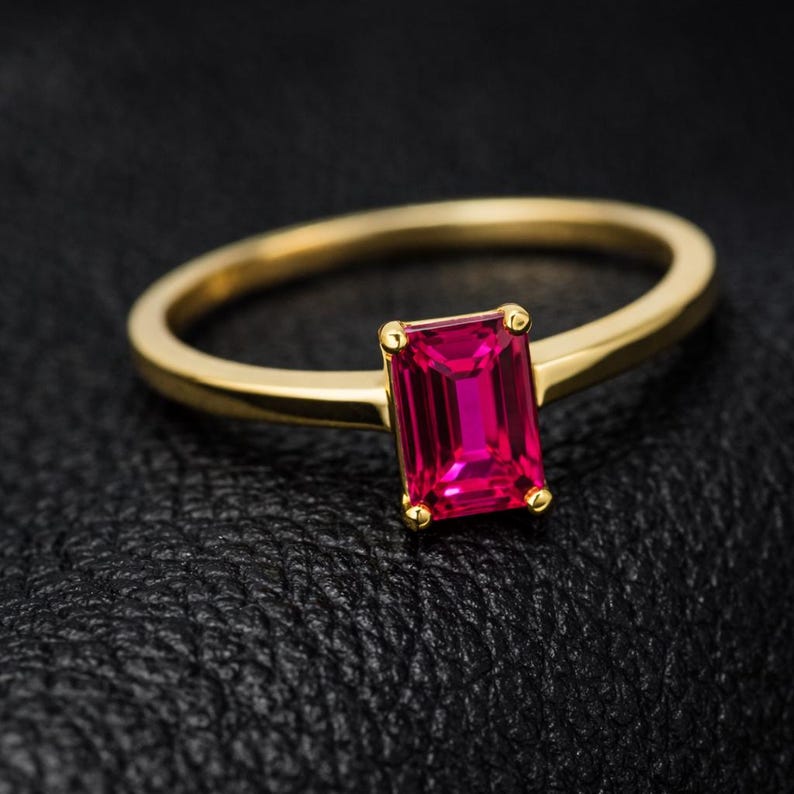 May include: A gold ring featuring a rectangular, vibrant pink gemstone. The ring's band is a simple, polished gold, and the gemstone is set with four small gold prongs. The ring is displayed against a dark, textured background.