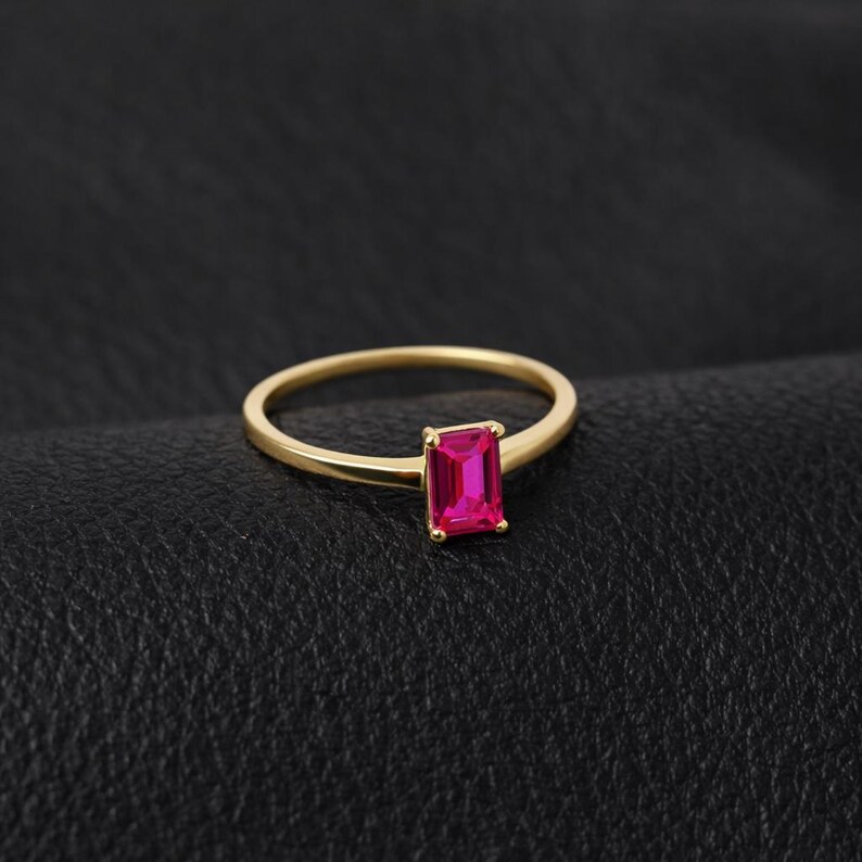 May include: A gold-colored ring with a rectangular, bright pink gemstone. The ring band is thin and simple, and the gemstone is set in a classic, minimalist style. The ring is displayed on a black surface.