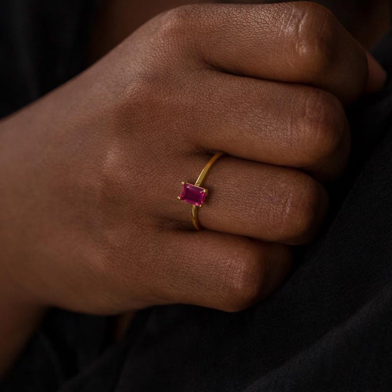May include: A gold ring featuring a rectangular, deep red gemstone. The ring is set on a slender gold band, and the gemstone is the focal point. The ring is shown on a hand with dark skin, against a dark background.