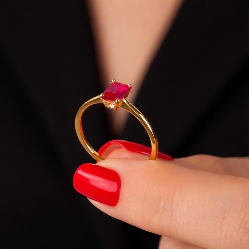 May include: A gold ring with a rectangular, bright red gemstone. The ring's band is thin and polished, and the gemstone is set in a four-prong setting. The ring is held up against a black background.