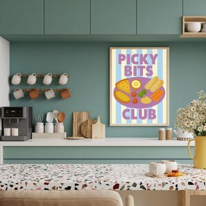Könnte beinhalten: Ein gerahmter Druck mit dem Text "PICKY BITS CLUB" und einer Lebensmittelillustration. Der Druck hängt an einer türkisfarbenen Wand über einer weißen Arbeitsplatte. Eine gelbe Kanne mit Blumen steht auf der Theke.