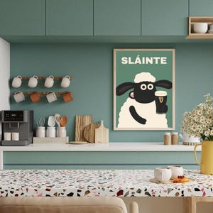 May include: A framed poster with the word "SL&Aacute;INTE" and a cartoon sheep holding a pint of stout. The poster is displayed on a teal wall in a kitchen setting. The kitchen also features a coffee maker, mugs, and a yellow pitcher with flowers.