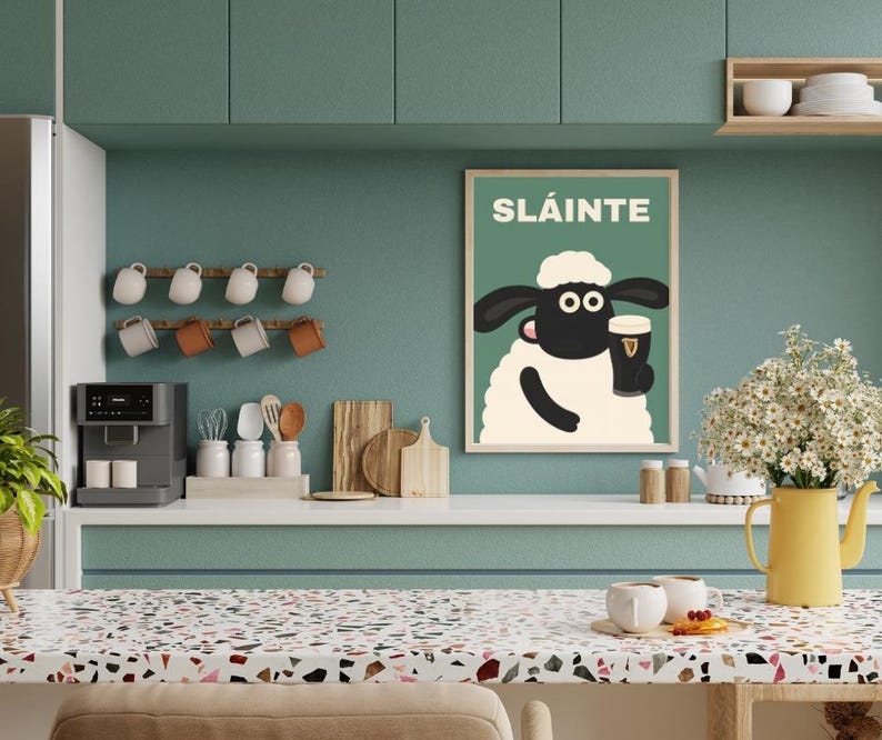 May include: A framed poster with the word "SL&Aacute;INTE" and a cartoon sheep holding a pint of stout. The poster is displayed on a teal wall in a kitchen setting. The kitchen also features a coffee maker, mugs, and a yellow pitcher with flowers.