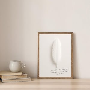 May include: A framed print with a large white feather on a white background. The wooden frame surrounds the artwork, and the text "Live a life so full, when you meet again you can tell them all about it" is written in cursive. A stack of books and a mug are on the left.
