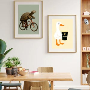 Puede incluir: Dos impresiones art&iacute;sticas enmarcadas. Una presenta una tortuga montando una bicicleta roja sobre un fondo verde. La otra muestra un pato blanco de dibujos animados sosteniendo una pinta de Guinness sobre un fondo amarillo. Ambas en marcos dorados.