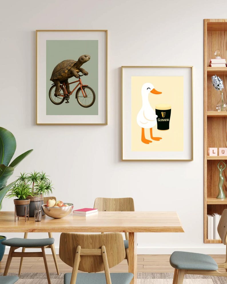 Puede incluir: Dos impresiones art&iacute;sticas enmarcadas. Una presenta una tortuga montando una bicicleta roja sobre un fondo verde. La otra muestra un pato blanco de dibujos animados sosteniendo una pinta de Guinness sobre un fondo amarillo. Ambas en marcos dorados.