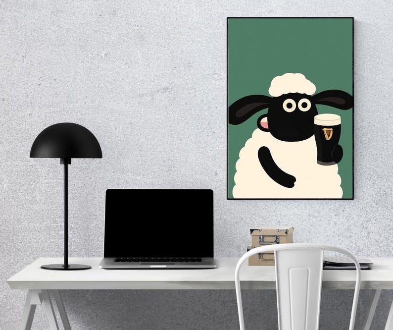 May include: A framed cartoon print featuring a sheep holding a pint of stout. The sheep has a white body, black face and ears, and is set against a green background. The pint glass has the Guinness logo. The print is displayed on a wall above a desk.
