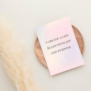 May include: A pastel-colored affirmation card with the text "I create a life filled with joy and purpose" sits on a wooden plate. A fluffy, beige decorative plant is to the left of the card.