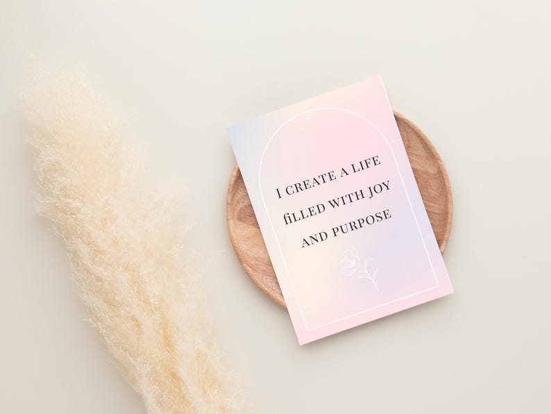 May include: A pastel-colored affirmation card with the text "I create a life filled with joy and purpose" sits on a wooden plate. A fluffy, beige decorative plant is to the left of the card.