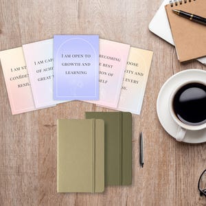 May include: An overhead shot of a wooden desk with motivational cards, journals, and a coffee cup. The cards have phrases like "I am open to growth and learning." Two journals are olive and tan. A pen, glasses, and laptop are also present.
