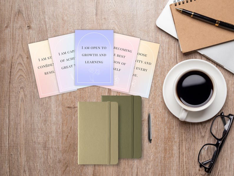 May include: An overhead shot of a wooden desk with motivational cards, journals, and a coffee cup. The cards have phrases like "I am open to growth and learning." Two journals are olive and tan. A pen, glasses, and laptop are also present.