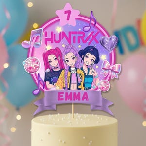 Peut inclure: Un décor de gâteau avec trois filles de style anime aux cheveux roses, violets et noirs. Le décor a un design circulaire rose et violet avec le texte "HUNTRAX" et le chiffre "7". Le nom "EMMA" est sur une bannière violette.