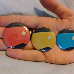 May include: Three round enamel pins with black and colored designs. The pins are red, yellow, and blue, each with a black border and a white and yellow emblem. The pins are held in a hand.