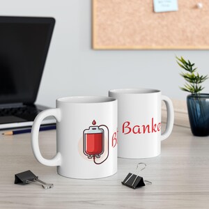 Peut inclure: Deux mugs en céramique blancs. L'un présente une illustration de poche de sang avec un liquide rouge et le texte « B ». L'autre mug porte le mot « Banker » en rouge. Les mugs sont sur une surface claire.