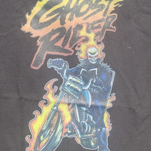 May include: Black t-shirt featuring a graphic of Ghost Rider on a motorcycle, engulfed in flames. The text "Ghost Rider" is displayed above the character in a fiery font, with a red outline.