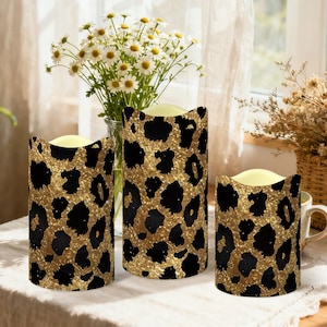May include: Three battery-operated pillar candles with a gold and black leopard print pattern. The candles are of varying heights and are placed on a table. A vase of white flowers and a basket of dried flowers are in the background.