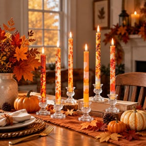 May include: Six tall taper candles with autumn-themed patterns, including orange, red, and yellow maple leaf prints and solid orange, are lit and placed in clear, textured glass candle holders on a table decorated with pumpkins and leaves.