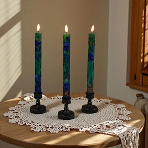 May include: Three lit, tall, green and blue floral patterned candles sit in black holders on a round table. The candles are on a white, crocheted doily. The table is made of wood.