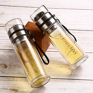 May include: Two clear glass tea infuser bottles with stainless steel lids and black straps. One bottle has a sunflower design, the other has the word "SLIGHTAIS" printed on it. Both bottles are filled with tea and sit on a wooden surface.
