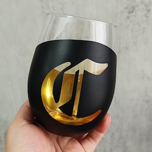 Puede incluir: Una copa de vino sin pie con base negra y diseño de letra "C" dorada. La parte superior de la copa es transparente. La copa se sostiene en una mano sobre un fondo neutro.