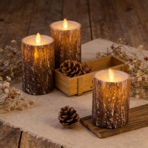 May include: Three battery-operated, cylindrical candles with a textured, brown bark-like exterior. Each candle has a warm, flickering LED light at the top, resembling a flame. A small wooden crate with pinecones sits between the candles, creating a rustic, cozy ambiance.