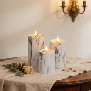 May include: Three marble-patterned pillar candles of varying heights with flickering LED flames. The candles are white with gray veining, creating a natural stone appearance. They sit on a wooden table with a neutral-colored cloth, alongside dried flowers.