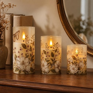 May include: Three cylindrical LED candles of varying heights, each featuring a cream-colored wax surface decorated with botanical illustrations of white flowers, green leaves, and butterflies, topped with a warm, glowing artificial flame.