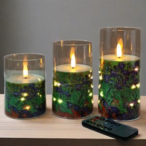 Op de afbeelding: Set van drie vlamloze LED-kaarsen in glazen cilinders. De kaarsen hebben een bloemenontwerp met groene, blauwe en rode tinten en zijn omwikkeld met lichtslingers. Een afstandsbediening ligt voor de kaarsen.
