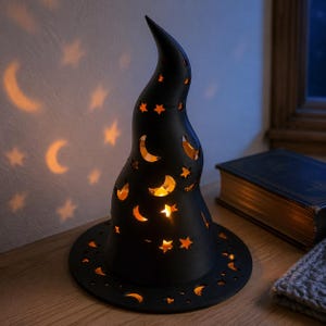 May include: A black witch hat-shaped lamp with cutouts in the form of stars and crescent moons. The lamp emits a warm orange glow from within, casting star and moon-shaped light patterns onto the nearby wall surface behind the object.