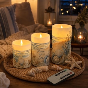 May include: Three cylindrical LED candles with a beach theme, featuring illustrations of seashells, starfish, and ocean waves. The centre candle displays the text "Find Peace by the Sea." A small remote control and loose seashells sit on a woven tray.