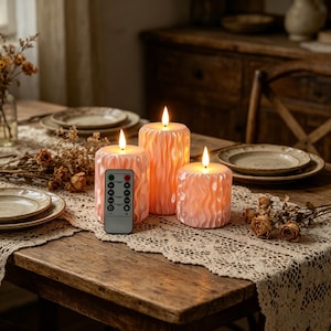 May include: Three peach-colored LED pillar candles with a wavy texture are displayed on a wooden table. A remote control lies near the candles. The candles are lit, and the scene is set on a table with plates and dried flowers.