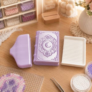 May include: A light purple plastic bead tray shaped like a vintage book with a crescent moon emblem on the cover, accompanied by a matching purple funnel and a white rectangular grooved sorting tray for organizing small craft beads.