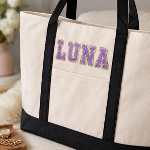 Op de afbeelding: Een crèmekleurige canvas draagtas met zwarte stoffen handvatten en een zwarte onderkant. De voorzijde van de tas toont de naam LUNA in paarse, textuurrijke letters in varsity-stijl met een gouden glitteromlijning boven een open zak.