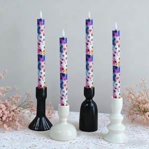 Może przedstawiać: Cztery dekoracyjne świece z kolorowym wzorem łap. Świece umieszczone są w czarno-białych świecznikach. Świece mają białą podstawę i kolorowy wzór łap w kolorze różowym, niebieskim i żółtym.