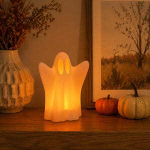 May include: A white, translucent ghost-shaped lamp emitting a warm, soft orange glow from within. The lamp features two oval-shaped eye cutouts and a smooth, rounded silhouette, standing upright as a decorative light fixture for seasonal decor.